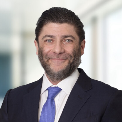 A man with short dark hair and a beard, wearing a dark suit, white shirt, and blue tie, stands in front of a blurred office background, representing chicago lawyers experienced in litigation support.