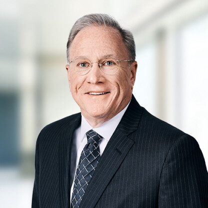 A middle-aged man with gray hair and glasses wearing a dark pinstripe suit, white shirt, and patterned tie stands in a blurred corporate law office setting.