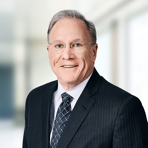 A middle-aged man with gray hair and glasses wearing a dark pinstripe suit, white shirt, and patterned tie stands in a blurred corporate law office setting.
