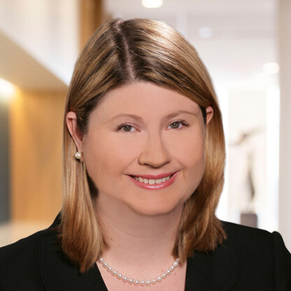 A woman with straight, shoulder-length blonde hair, wearing a black blazer, pearl necklace, and pearl earrings, smiles at the camera in a brightly lit law offices hallway known for supporting Chicago lawyers.