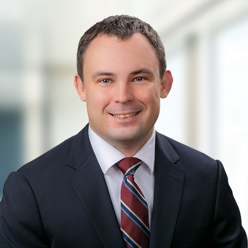 Man in a dark suit, white shirt, and striped tie, smiling at the camera in a bright, blurred law offices setting—an approachable professional experienced in intellectual property law.