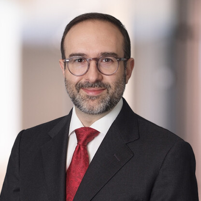 A man with short dark hair, glasses, and a beard, wearing a dark suit, white shirt, and red tie, stands in front of a blurred indoor background typical of modern law offices.