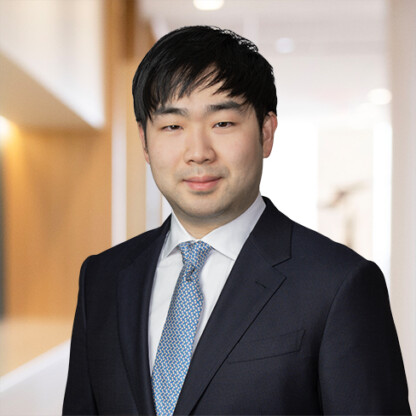 A man in a dark suit and patterned tie stands in a well-lit, modern law offices hallway, looking at the camera with a neutral expression.