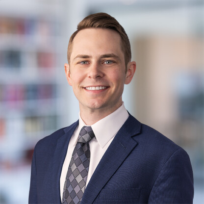 A man in a blue suit, white shirt, and patterned tie smiles at the camera in a blurred corporate law office setting.
