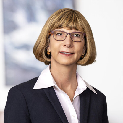 A woman with short blonde hair, glasses, and gold hoop earrings, wearing a white collared shirt and a dark blazer, poses in front of a blurred background typical of corporate law offices.