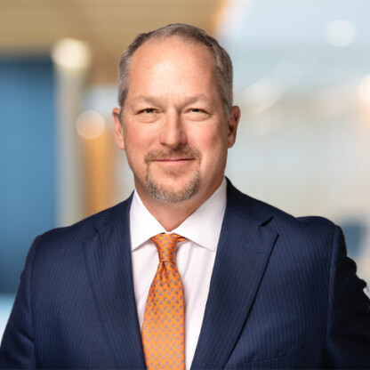 A middle-aged man wearing a blue pinstripe suit, white shirt, and orange patterned tie stands confidently in a modern corporate law office environment.
