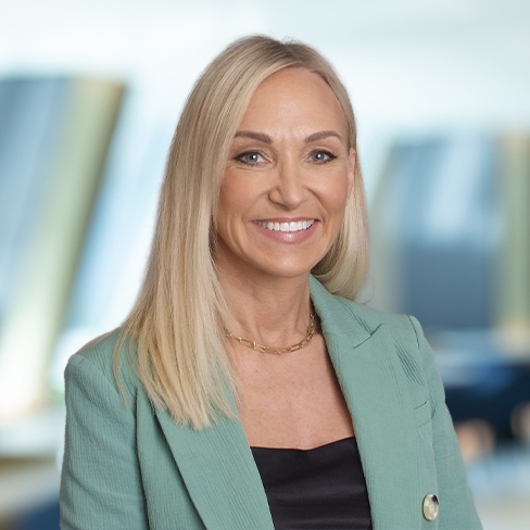A woman with straight blonde hair, wearing a light green blazer and black top, stands smiling in a modern, blurred law offices setting.