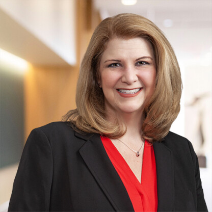 Woman with shoulder-length blonde hair wearing a black blazer and red blouse, smiling, standing in a brightly lit corporate law office among fellow Chicago lawyers.