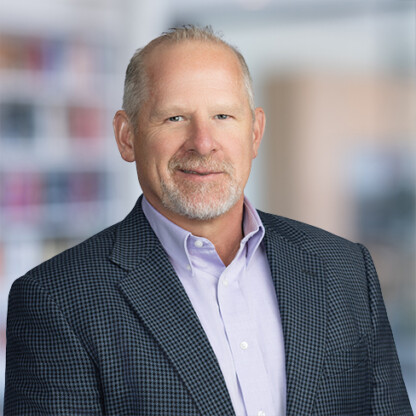 Middle-aged man with short gray hair and a trimmed beard, wearing a light purple shirt and dark checkered blazer, standing in an indoor corporate law office with blurred bookshelves in the background.