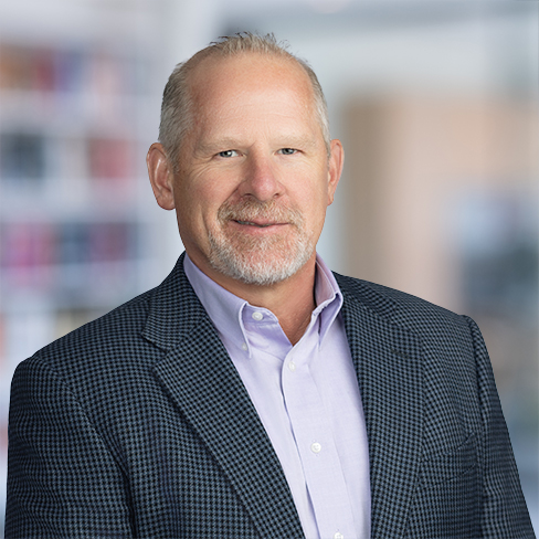 Middle-aged man with short gray hair and a trimmed beard, wearing a light purple shirt and dark checkered blazer, standing in an indoor corporate law office with blurred bookshelves in the background.