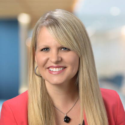A woman with long blonde hair, wearing a red blazer, necklace, and large hoop earrings, smiles in a modern law office setting, representing chicago lawyers with expertise in intellectual property law.