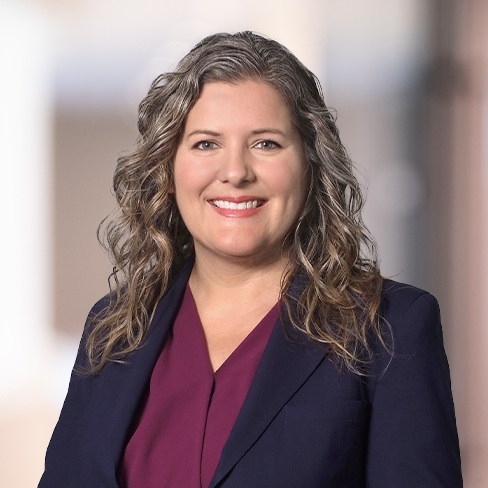 Woman with wavy, shoulder-length gray hair wearing a dark blazer over a burgundy top, smiling at the camera—a confident presence among leading lawyers in Chicago, specializing in intellectual property law. Blurred indoor background.