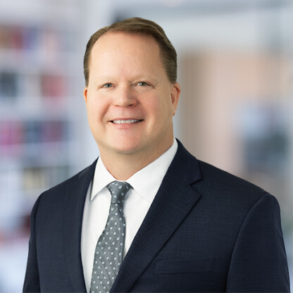 A man in a dark suit, white shirt, and patterned tie smiles while standing in a blurred office setting, representing litigation support for lawyers in Chicago.