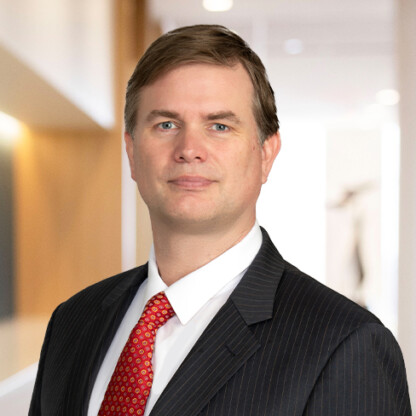 A man in a dark pinstripe suit, white shirt, and red tie stands in a modern, well-lit corporate law office hallway, looking directly at the camera.