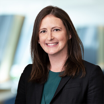A woman with straight brown hair wearing a dark blazer and green top, smiling in a corporate law office setting with blurred background.