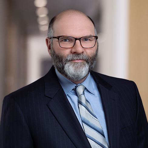 A middle-aged man with a beard and glasses, wearing a dark pinstripe suit, blue shirt, and striped tie, stands in a hallway—typical of lawyers in Chicago specializing in intellectual property law.