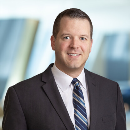 A man in a dark suit, white shirt, and striped tie smiles at the camera against a blurred corporate law office background.