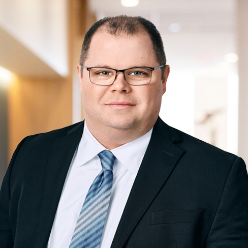 A man wearing glasses, a dark suit, white shirt, and striped tie stands in a brightly lit office hallway, exuding the professionalism often seen among Chicago lawyers specializing in intellectual property law.