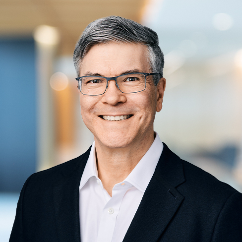 A man with short gray hair and glasses, wearing a dark suit jacket and white shirt, smiles at the camera in a modern corporate law office setting.