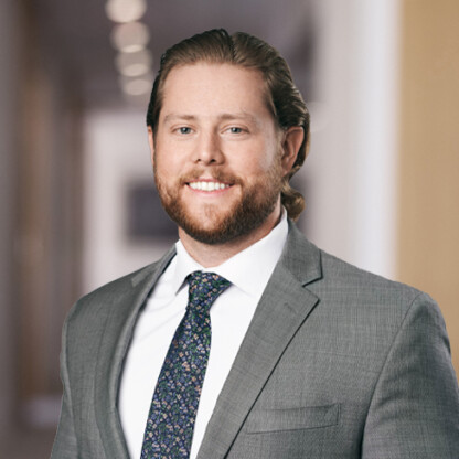 A man with long, light brown hair and a beard, wearing a gray suit, white shirt, and patterned tie, stands in a blurred hallway of a corporate law office.