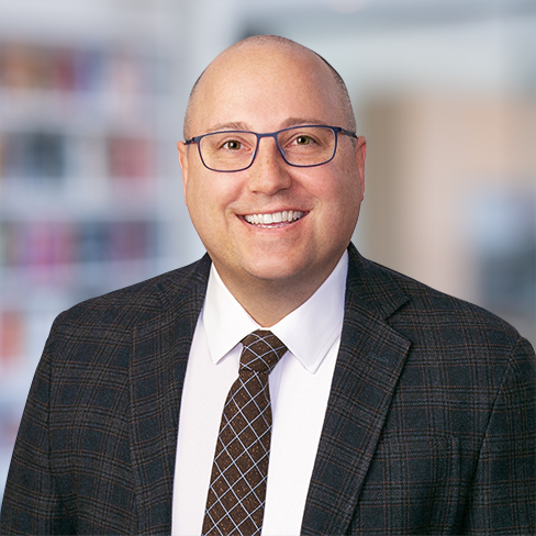 Smiling man in glasses wearing a checked suit jacket, white shirt, and patterned tie, standing in what appears to be law offices or a library—an ideal image for chicago lawyers specializing in intellectual property law.