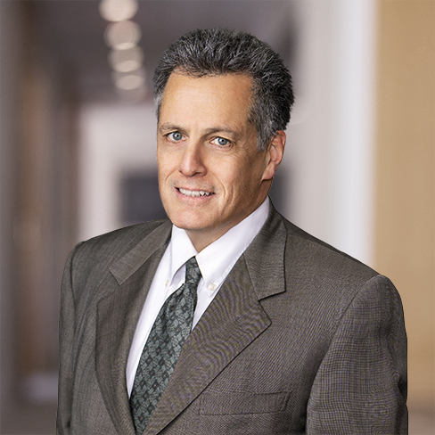 A middle-aged man in a gray suit, white shirt, and patterned tie stands in a blurred law office hallway, looking at the camera with a neutral expression, suggesting expertise in intellectual property law.
