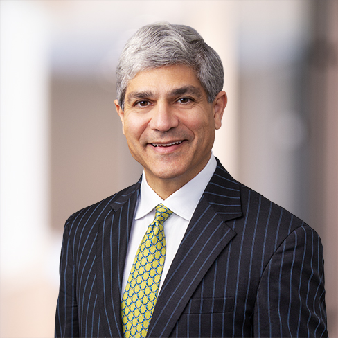 A man with short gray hair, wearing a dark pinstripe suit, white shirt, and patterned yellow tie, stands in a blurred indoor setting often seen in law offices of top Chicago lawyers.