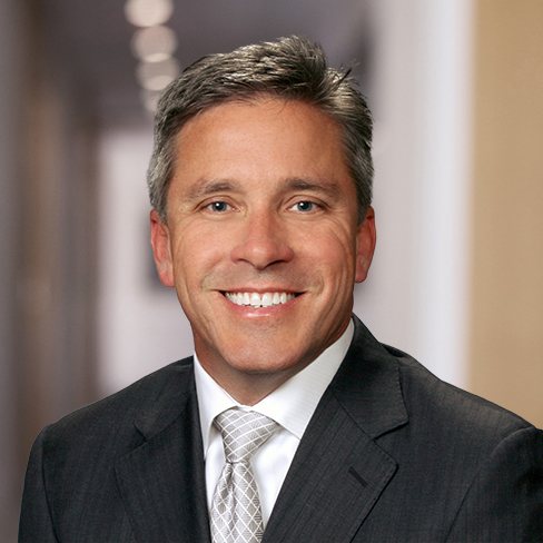 Smiling man in a suit and tie standing in a blurred corporate law office hallway.