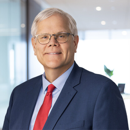 Man with short gray hair and glasses, wearing a navy suit, light blue shirt, and red tie, standing in a modern corporate law office.