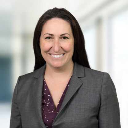 A woman with long dark hair wearing a gray blazer and maroon patterned blouse smiles at the camera, standing in a bright, blurred indoor setting—representing experienced lawyers in Chicago.