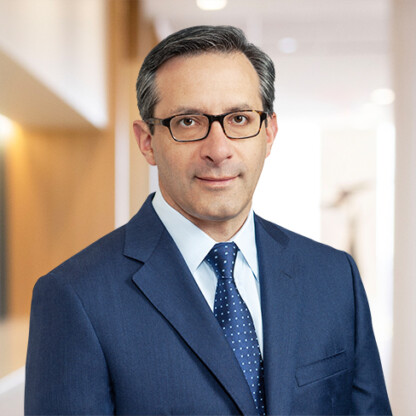A man wearing glasses and a blue suit with a polka dot tie stands in a brightly lit corporate law office hallway.