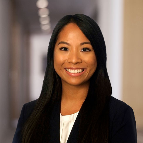 A woman with long black hair, wearing a dark blazer and white top, smiles at the camera in a hallway with blurred lights, capturing the professional atmosphere of a corporate law office.