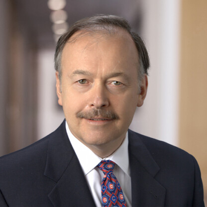 A middle-aged man with a mustache and short hair wearing a dark suit, white shirt, and patterned tie, stands in a hallway with blurred lights in the background, embodying professionalism often seen in corporate law office environments.