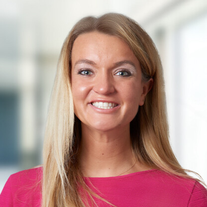 Woman with long blonde hair, wearing a pink top, smiling and standing in a bright, blurred indoor setting at one of Chicago’s premier law offices specializing in intellectual property law.