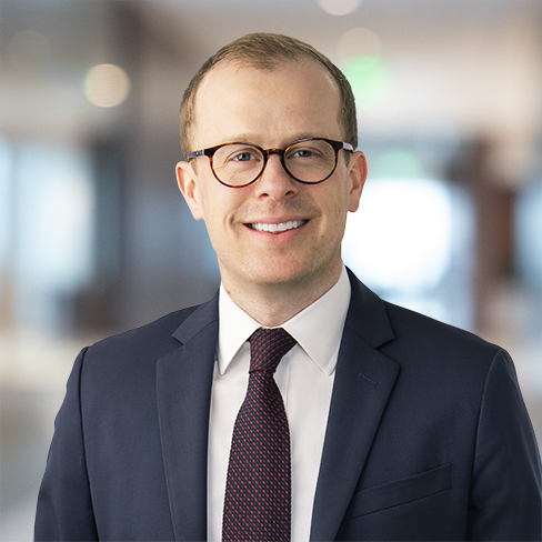 A man in a suit, tie, and glasses stands in a modern corporate law office, smiling at the camera.