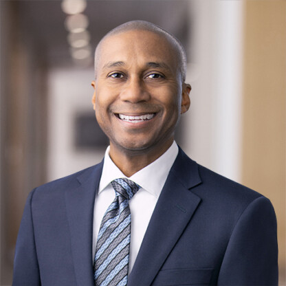 A man in a dark suit and patterned tie smiles at the camera while standing in a corporate law office hallway with blurred lights in the background.
