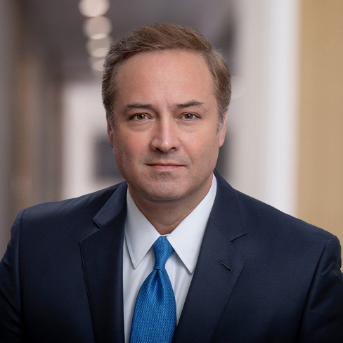 A man in a suit and blue tie poses for a formal portrait in the hallway of law offices, with blurred lights in the background, reflecting his expertise in litigation support.