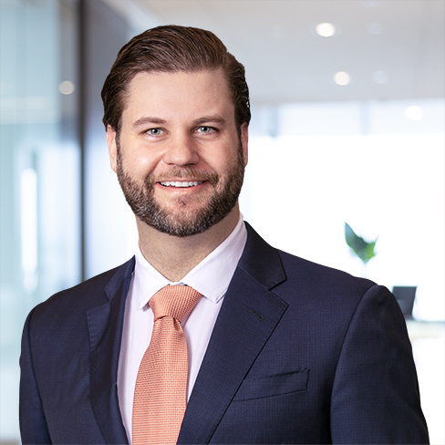 A man in a navy suit, white shirt, and peach tie smiles while standing in a modern corporate law office with glass walls and a blurred background.