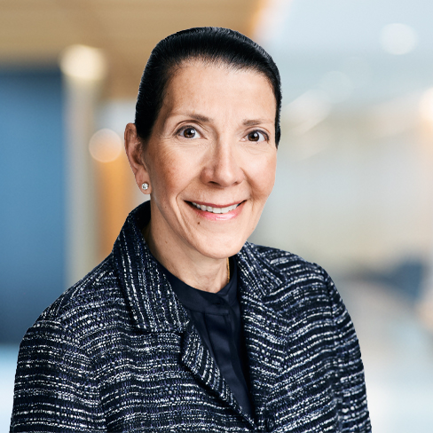 A woman with dark hair pulled back, wearing a dark tweed blazer and stud earrings, smiles at the camera in an office setting, reflecting the professionalism found in Chicago lawyers and law offices.