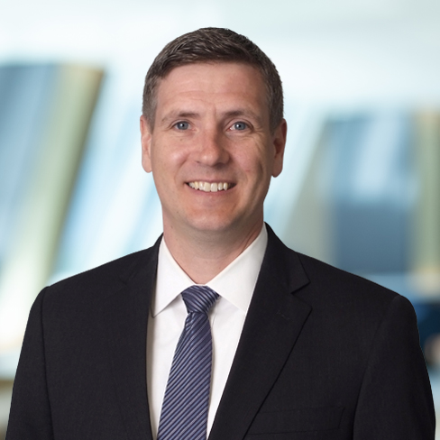 A man in a dark suit, white shirt, and striped tie smiles at the camera, standing in front of a blurred background with blue and beige tones—reflecting the professionalism of Chicago lawyers specializing in intellectual property law.