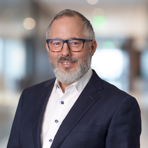A man with a gray beard and glasses, dressed in a dark suit and white shirt, stands in a blurred indoor setting—typical of seasoned Chicago lawyers experienced in litigation support.