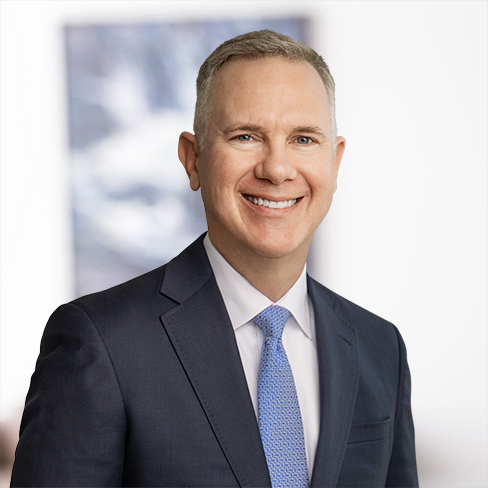 A man in a navy suit, light blue tie, and white shirt smiles while standing in front of a blurred corporate law office background.