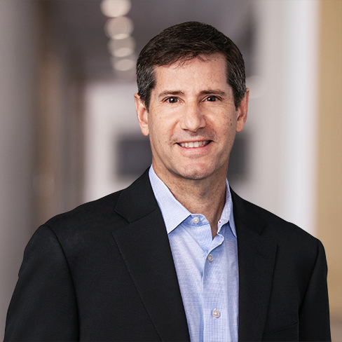 A man with short brown hair wearing a dark blazer and light blue dress shirt stands in the hallway of a corporate law office, with a blurred background suggesting a busy environment for lawyers in Chicago.