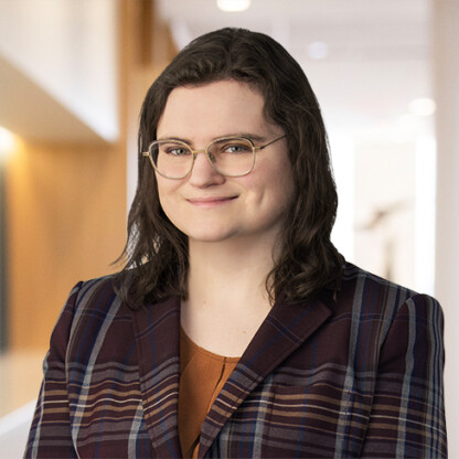 A person with shoulder-length brown hair and glasses, wearing a plaid blazer and orange top, stands indoors in a well-lit hallway of a corporate law office, reflecting the professional atmosphere of Chicago lawyers.