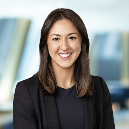 A woman with straight brown hair, wearing a black blazer and dark top, smiles at the camera against the blurred backdrop of a corporate law office.