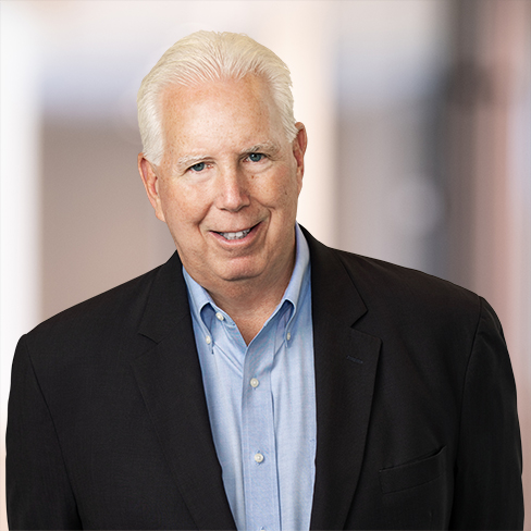 An older man with white hair, wearing a blue shirt and dark blazer, stands in front of a blurred indoor background, smiling at the camera—a confident professional among Chicago lawyers known for expertise in intellectual property law.