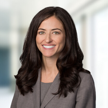 A woman with long brown hair, wearing a gray blazer and top, smiles at the camera in a brightly lit corporate law office in Chicago.