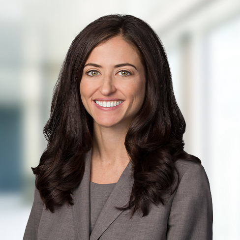 A woman with long brown hair, wearing a gray blazer and top, smiles at the camera in a brightly lit corporate law office in Chicago.
