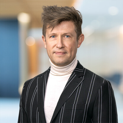 Man with short, styled brown hair wearing a black blazer with white pinstripes and a white turtleneck, standing in a brightly lit, modern corporate law office that offers litigation support.
