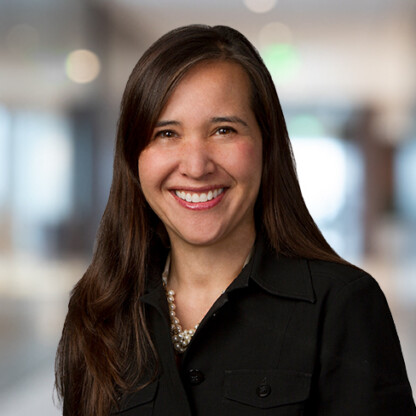 Woman with long brown hair, wearing a black jacket and pearl necklace, smiling in a blurred corporate law office, representing the professionalism of lawyers in Chicago.
